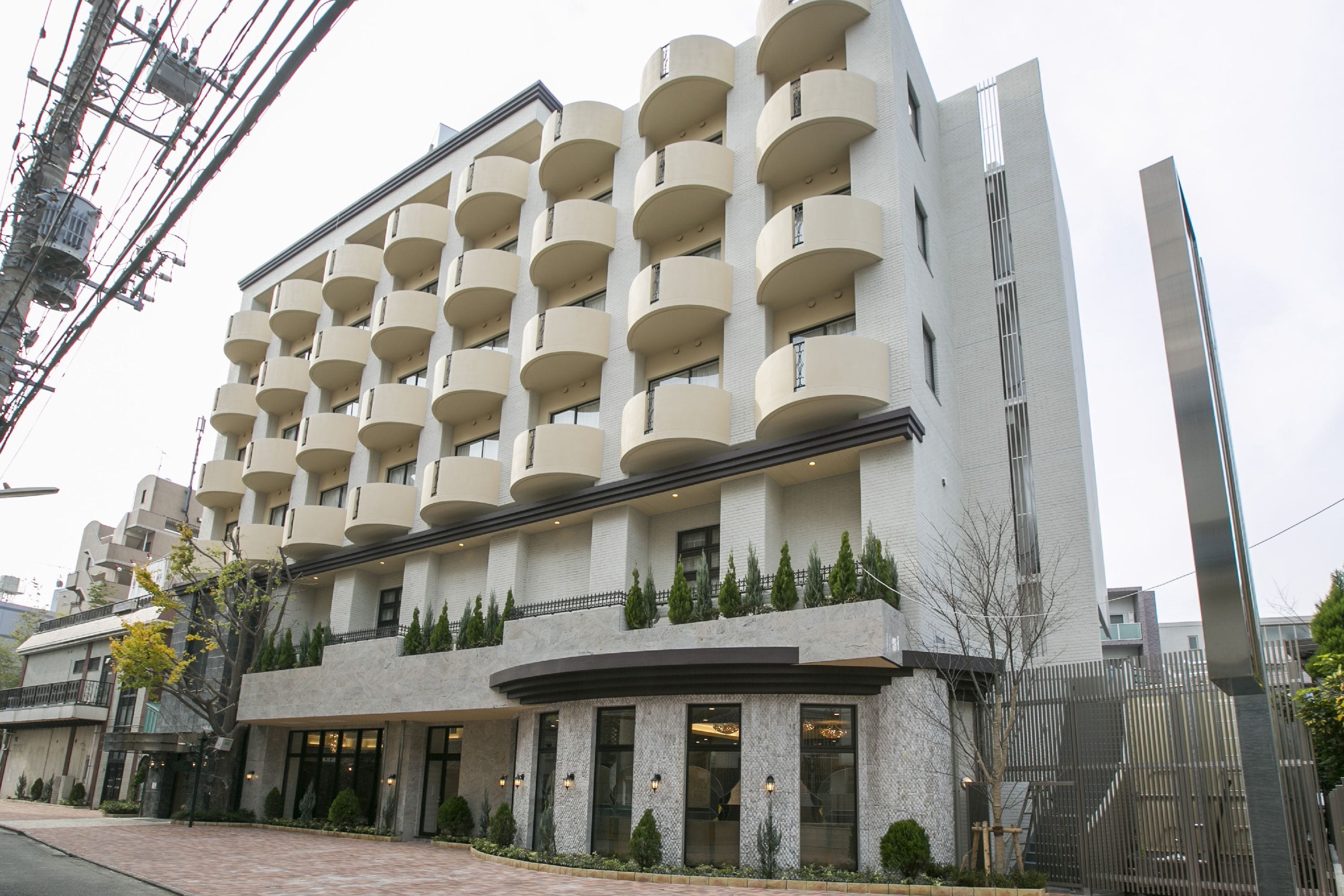 A multi-story hotel with a modern design, featuring balconies on each floor, a ground-level entrance, and a landscaped area with small trees.