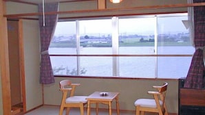 A room with a large window overlooking a body of water, two wooden chairs, a small table, and a wooden bench. - Ayame-ryokan (Itako)