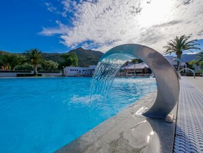 A swimming pool with a modern water feature, surrounded by palm trees and mountains in the background.
