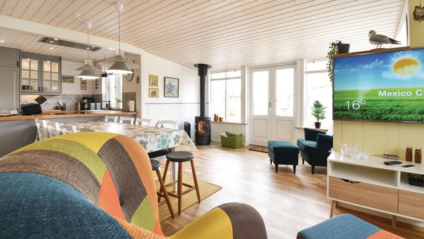 Un salon moderne avec un grand téléviseur diffusant un paysage, un coin salon confortable agrémenté de coussins colorés, une cuisine en arrière-plan et une cheminée.