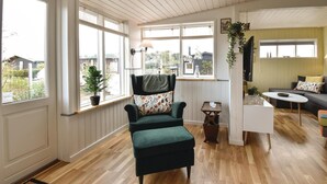 Un salon chaleureux avec un fauteuil vert, une petite table et un canapé. De grandes fenêtres laissent entrer la lumière naturelle et offrent une vue sur l’extérieur.