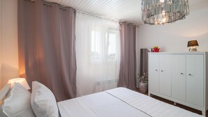 Een slaapkamer met een bed, witte gordijnen, een witte kast, een kroonluchter en een vaas met bloemen.