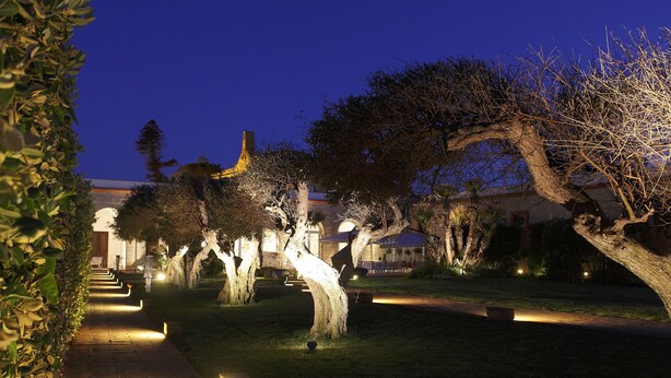 A nighttime view of a well-lit pathway lined with trees and a building in the background. - I Pretti Resort (Favignana)