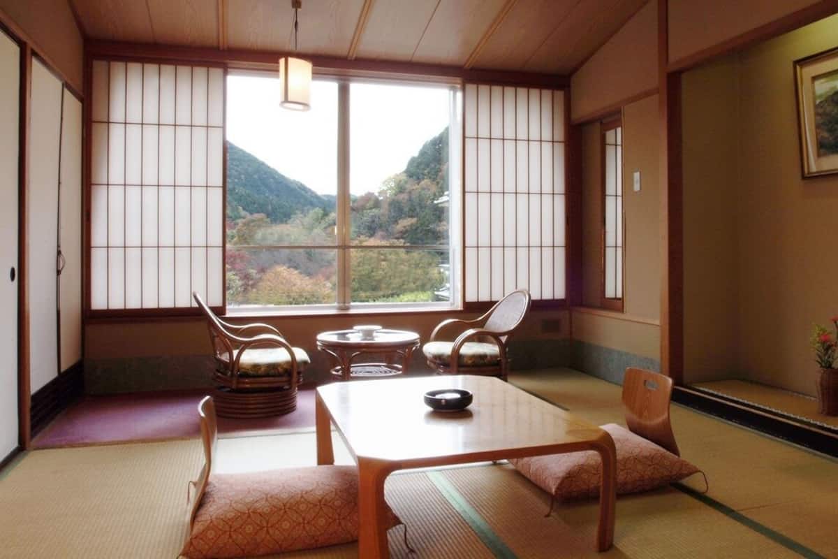 A traditional Japanese room with tatami flooring, a low table, chairs, and a view of a mountainous landscape through large windows.