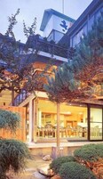 Edifício moderno com área de refeições visível através de grandes janelas de vidro, cercado por um jardim arborizado com árvores e arbustos.