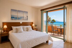 A hotel room with a bed, two bedside tables, a nightstand, a balcony with a table and chairs, and a view of the sea.