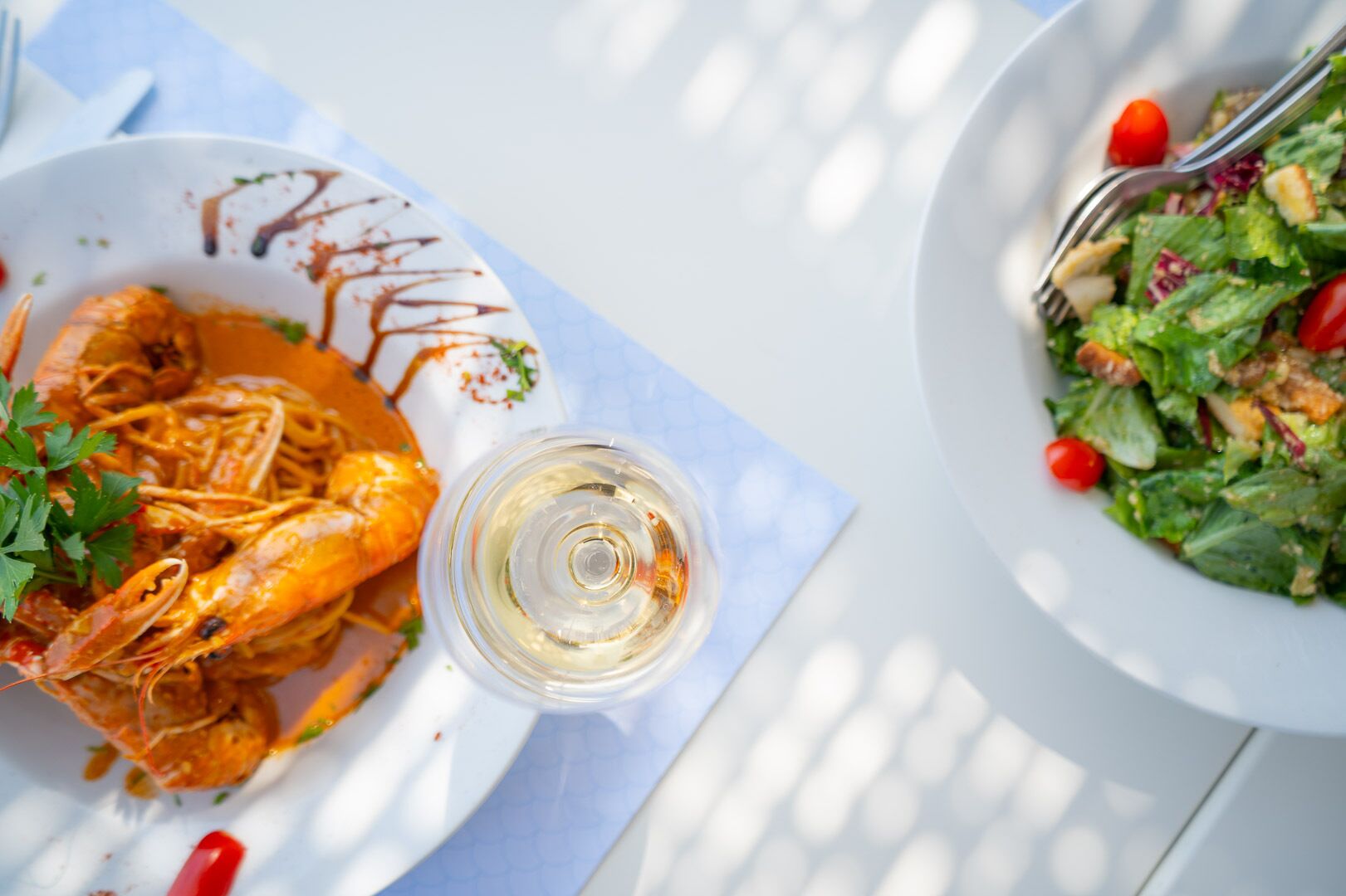 Un plato de camarones con una copa de vino blanco y una ensalada al lado, sobre una mesa blanca.