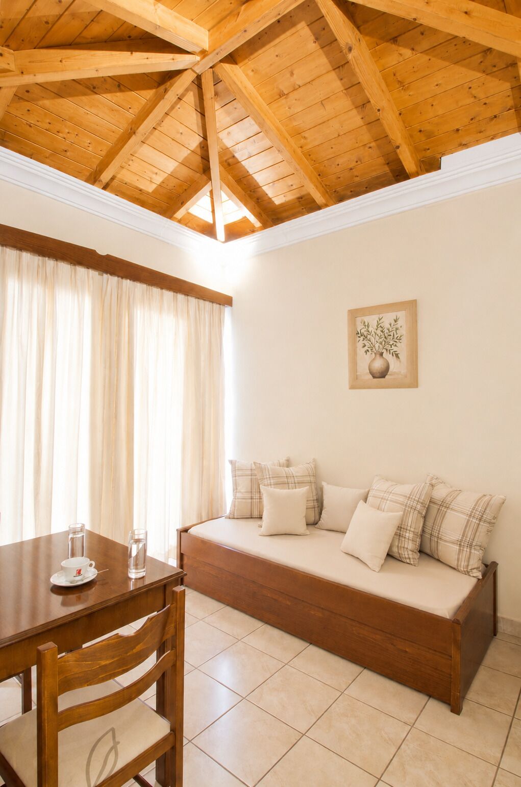 Una habitación con techo de madera, un sofá con cojines escoceses, una mesa de comedor con sillas y una planta en un marco en la pared.