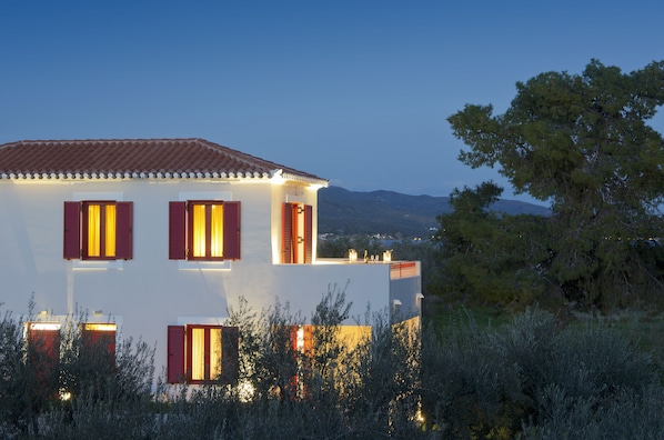 A two-story building with red shutters and a white facade, surrounded by vegetation and mountains in the background.