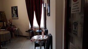 A dining area with a round table covered with a lace tablecloth, two black chairs, and a vase with flowers. A painting is on the wall, and a window with red curtains is visible. - Hotel Houston (Livorno)