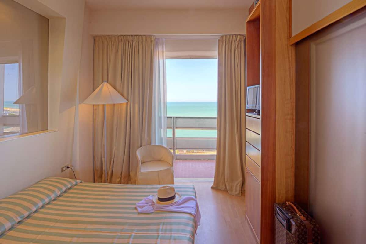 A hotel room with a bed, a chair, a TV, and a view of the sea.