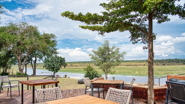 An outdoor deck with wooden tables and chairs, overlooking a body of water and grassy landscape. - Chobe River Camp (Ngoma)