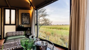 A room with a bed, a table with glasses and a metal cup, a painting on the wall, and a view of a grassy field through a window. - Chobe River Camp (Ngoma)