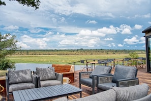 A deck with outdoor furniture overlooking a lake and a grassy landscape. - Chobe River Camp (Ngoma)