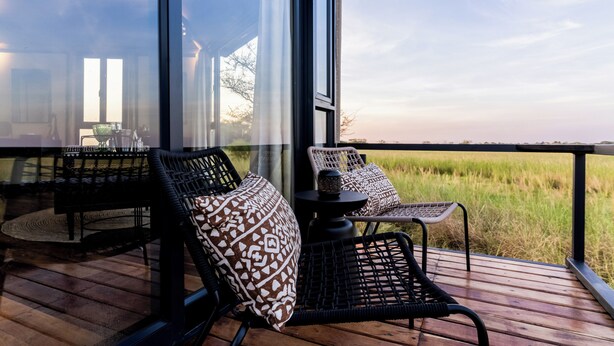 A deck with wicker chairs and a table, overlooking a grassy field. - Chobe River Camp (Ngoma)