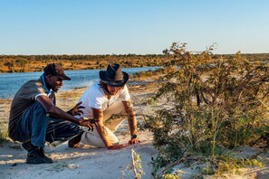 Two people on a sandy beach near water with sparse vegetation. - Chobe River Camp (Ngoma)
