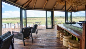 A spacious veranda with wooden flooring, a bar area with stools, and large glass doors leading to an outdoor view. - Chobe River Camp (Ngoma)