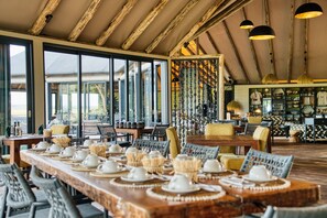 A restaurant with a long dining table set for a meal, wooden beams on the ceiling, and large windows. - Chobe River Camp (Ngoma)