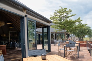 An outdoor dining area with wooden tables and chairs, large glass doors, and a view of the surrounding landscape.