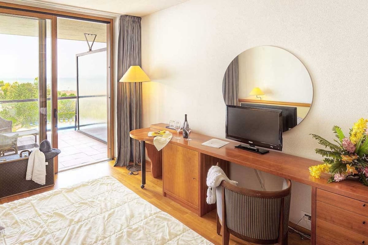 A hotel room with a bed, a desk with a television, a chair, a small table, and a balcony with a view of the sea.