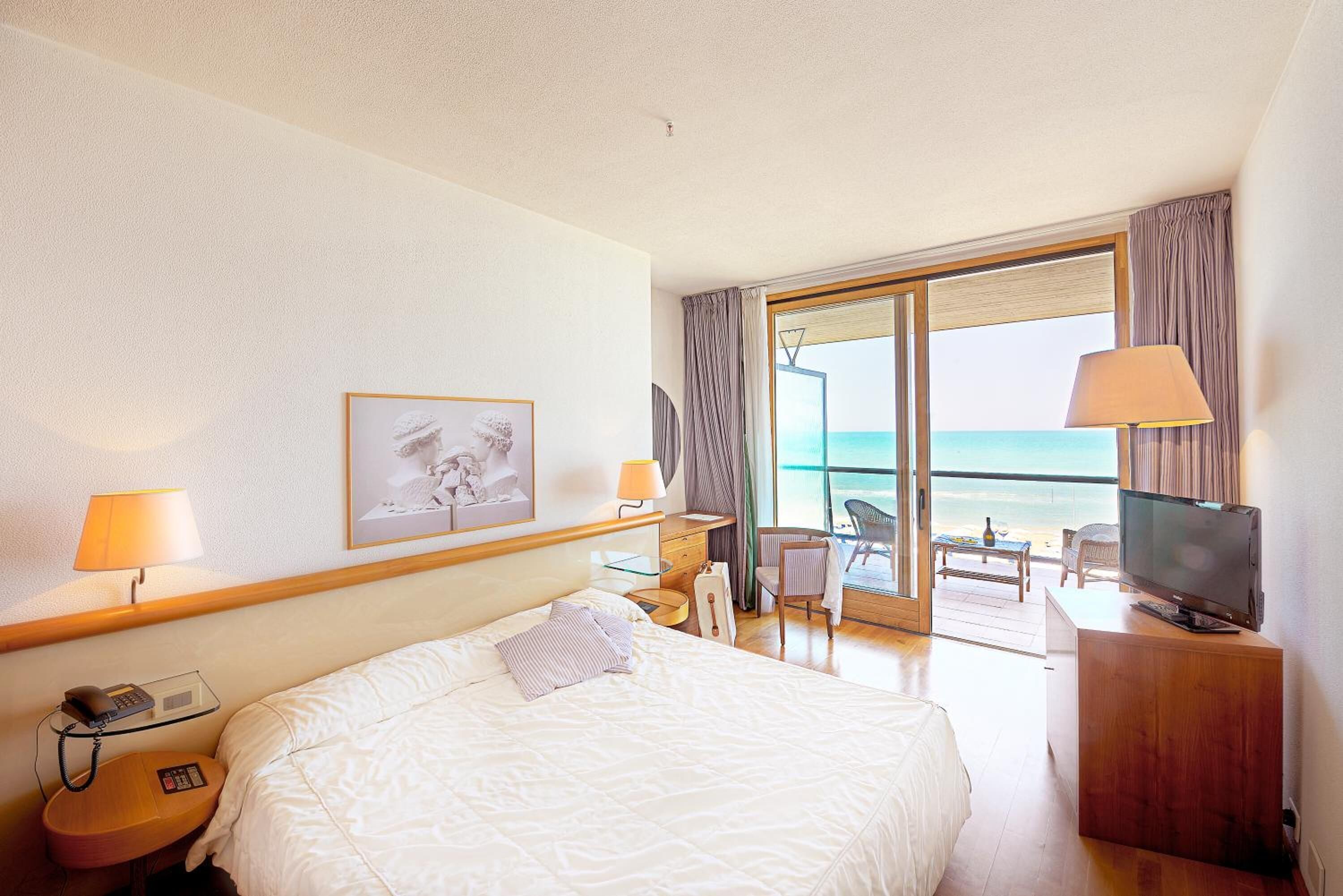 A hotel room with a bed, a desk, a chair, a television, and a view of the beach.