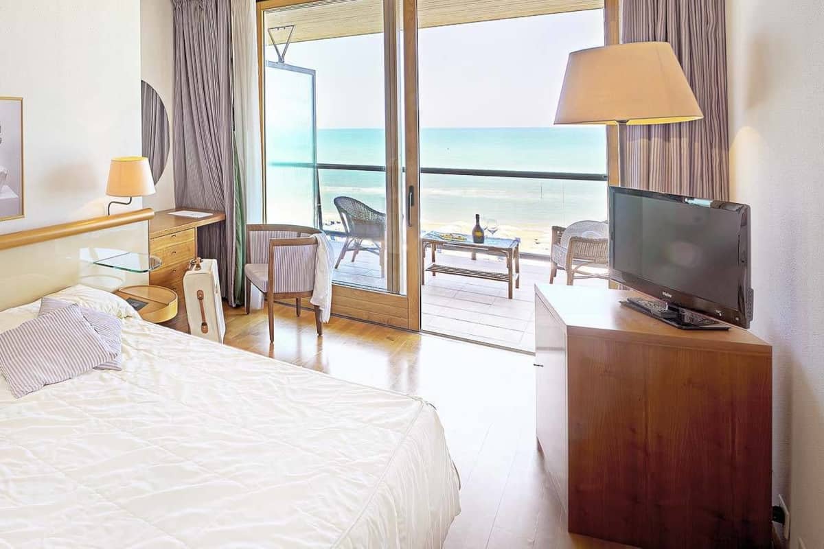 A hotel room with a bed, bedside lamps, a desk with a chair, a television, and a large window with a view of the beach.