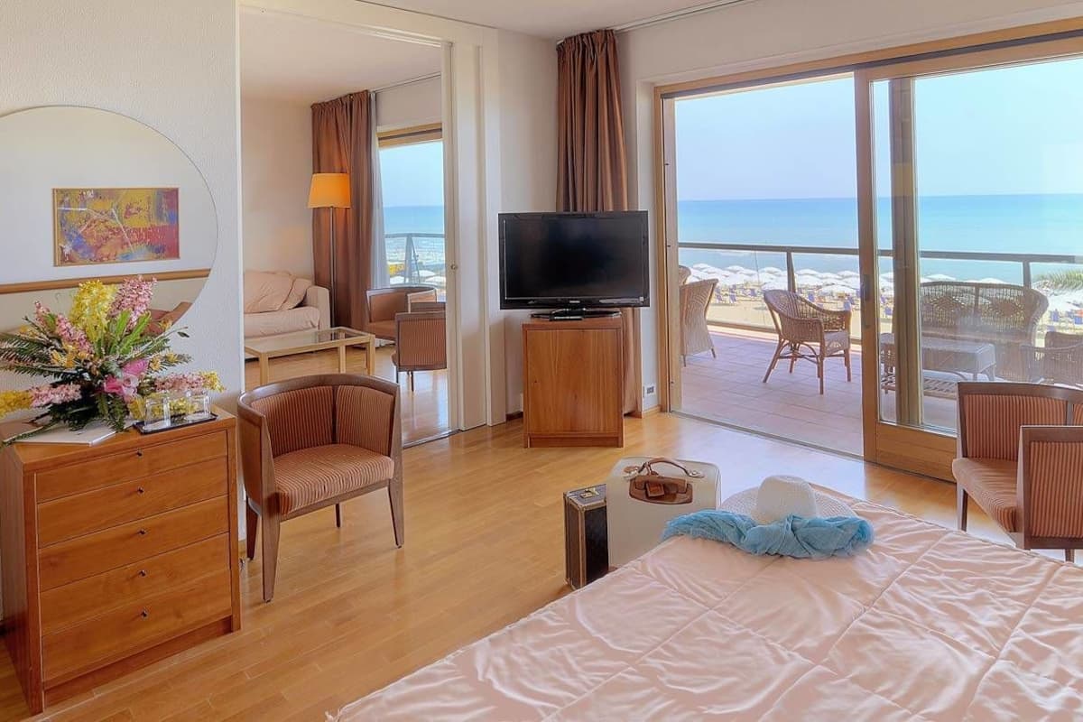 A hotel room with a bed, a TV, a desk, a chair, and a balcony with a view of the beach.