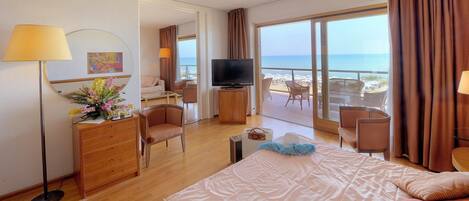 A hotel room with a bed, a TV, a desk, a chair, and a balcony with a view of the beach.