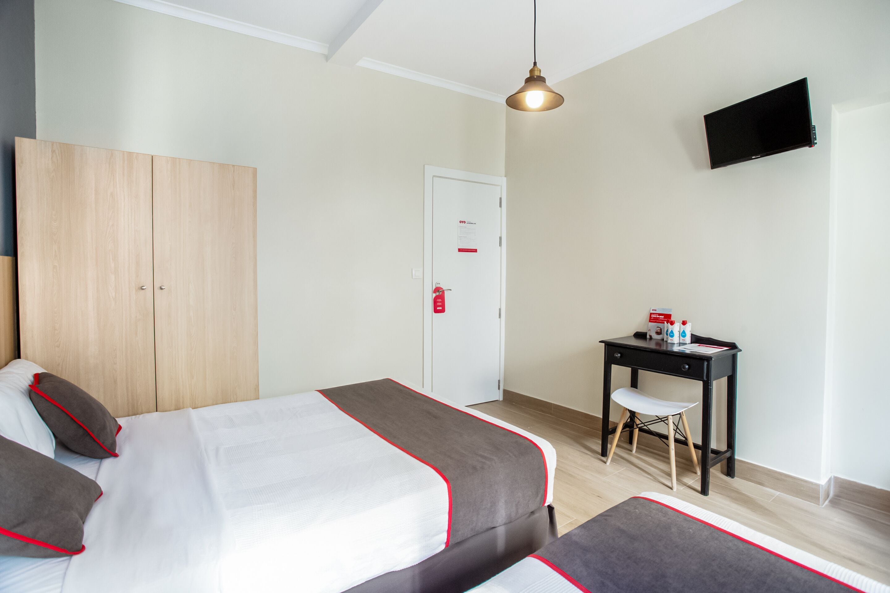 A hotel room with two beds, a desk, a chair, a TV, and a door with a fire alarm.