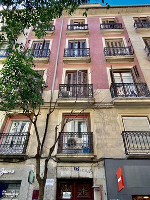 Un edificio de varios pisos con balcones, una tienda con un letrero y un árbol en primer plano.