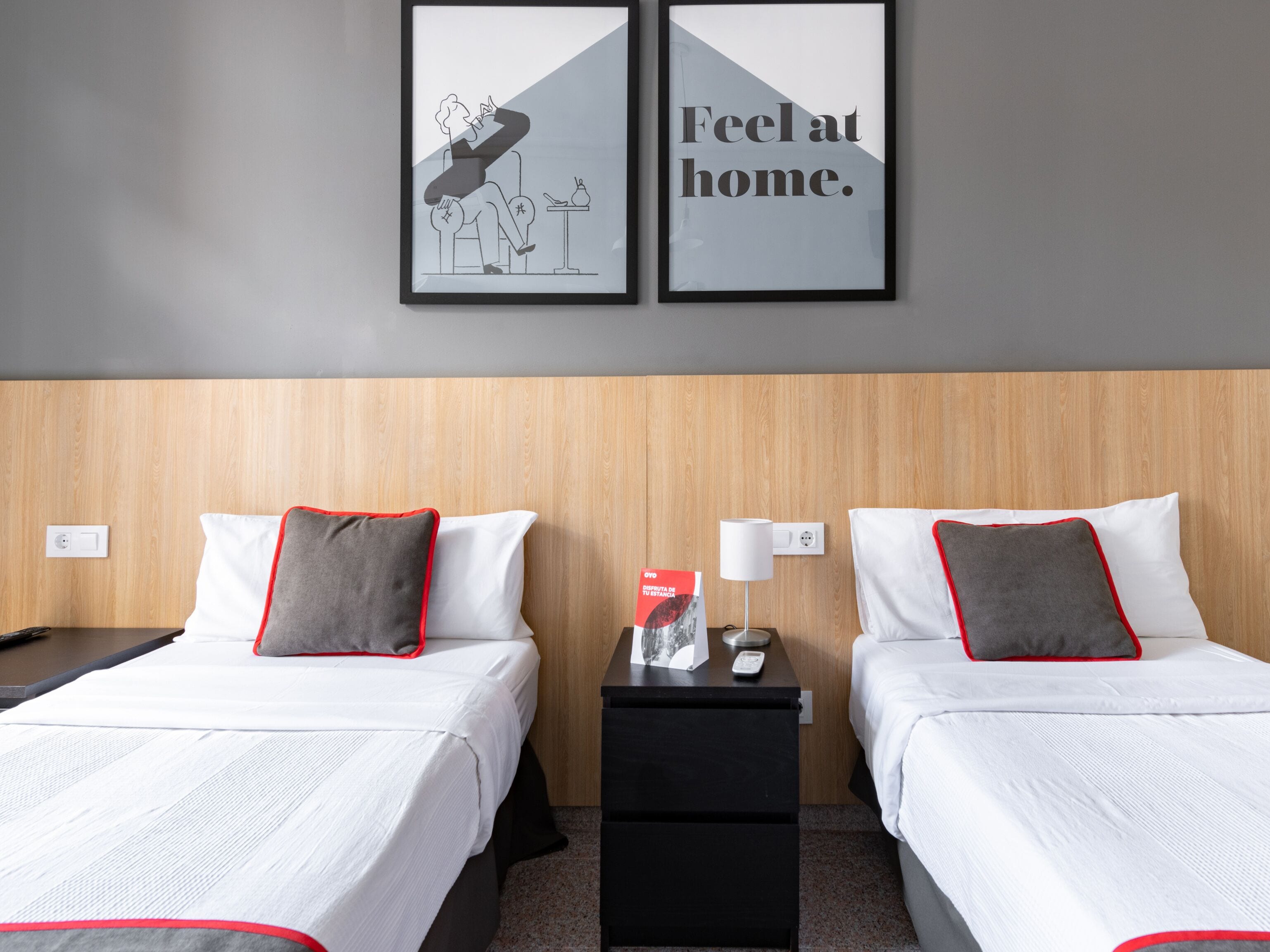 Habitación de hotel con dos camas, cada una con ropa de cama blanca y almohadillas con detalles rojos. Hay dos cuadros en la pared, uno que muestra a una persona sentada y otro con el texto "Feel at home".