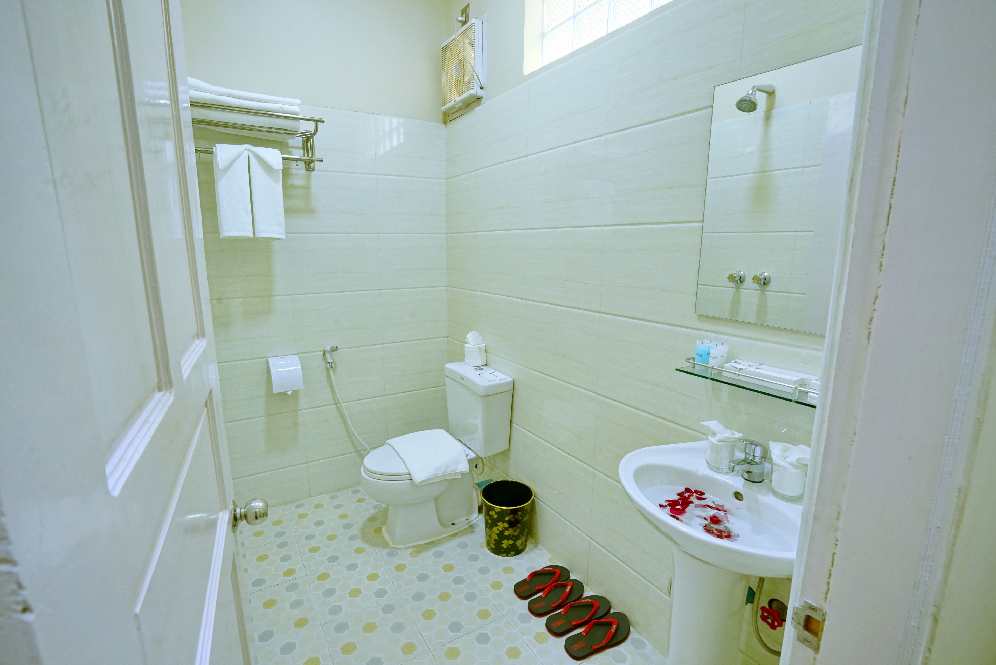 A bathroom with a toilet, sink, and shower. There are slippers on the floor and a trash can.