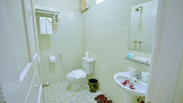 A bathroom with a toilet, sink, and shower. There are slippers on the floor and a trash can.