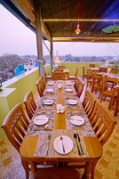 Una larga mesa de comedor dispuesta con platos, cubiertos y un florero central, rodeada de sillas de madera, bajo una zona cubierta con vistas a edificios y árboles.