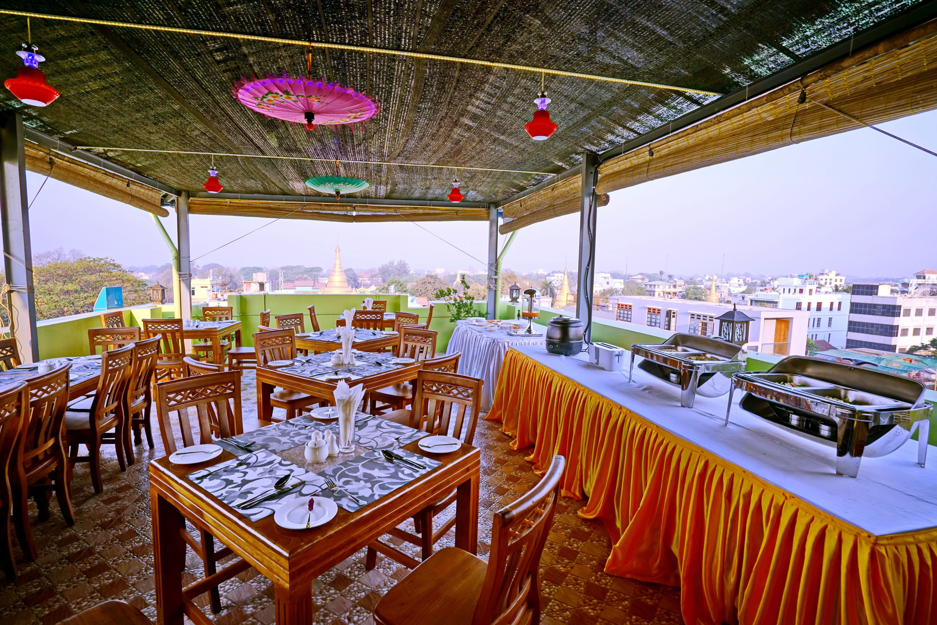 An outdoor dining area with tables set for meals, a buffet line, and a view of the cityscape in the background.