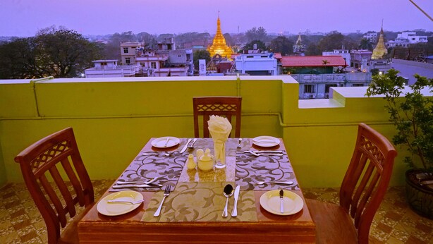 A dining table set with plates, cutlery, and a vase, overlooking a cityscape with buildings and a golden pagoda. - Hotel Shwe Phyo (Mandalay)