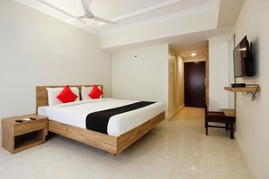 A hotel room with a wooden bed, a nightstand, a TV mounted on the wall, and a door leading to another room. - Royal Derby Suprabhat (Hyderabad)