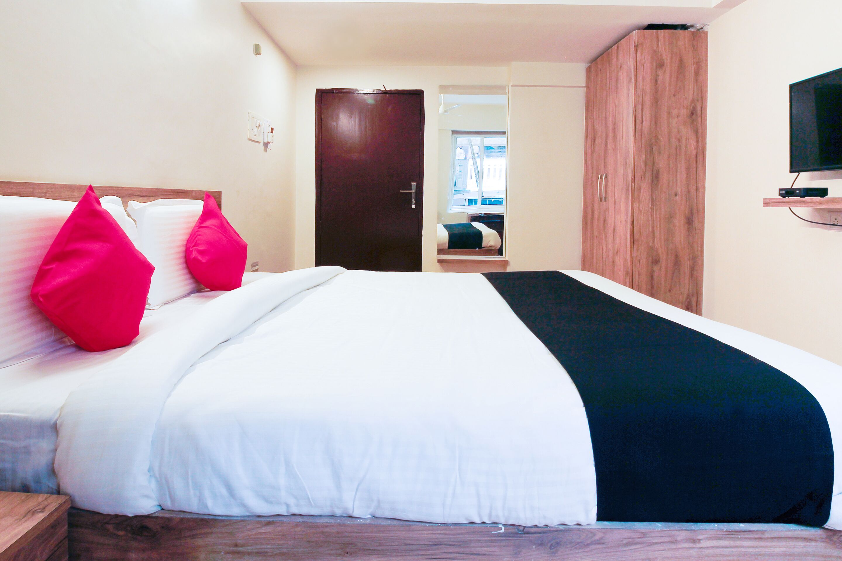 A hotel room with a bed, a wooden wardrobe, a television mounted on the wall, and a window.