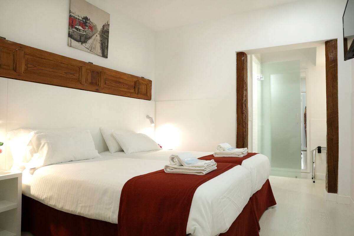 Habitación de hotel con dos camas, ropa de cama blanca, cabecero de madera, un cuadro en la pared y una puerta de cristal que da a un baño.