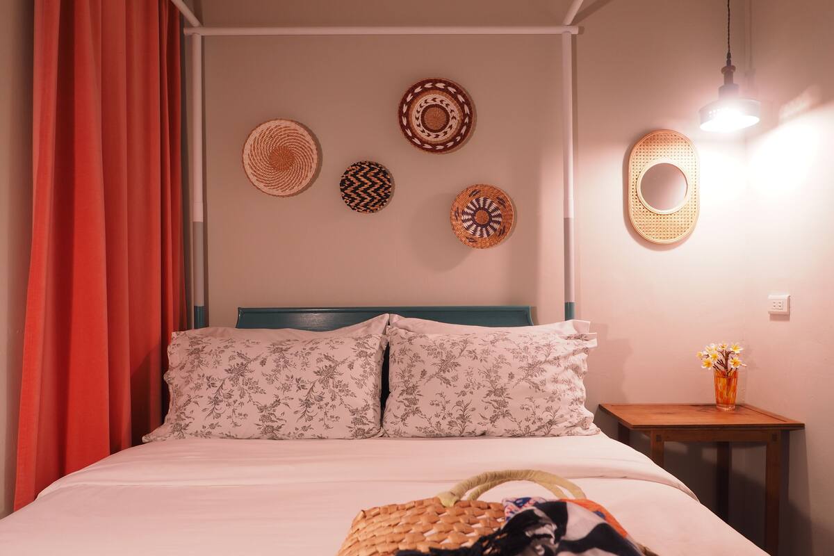 A neatly made bed with floral-patterned pillows, a woven basket on the bed, and a small table with a vase of flowers beside it.