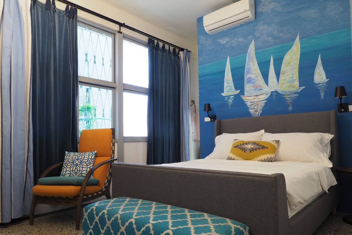 A hotel room with a bed, a chair, a window with curtains, and a wall painting of sailboats.