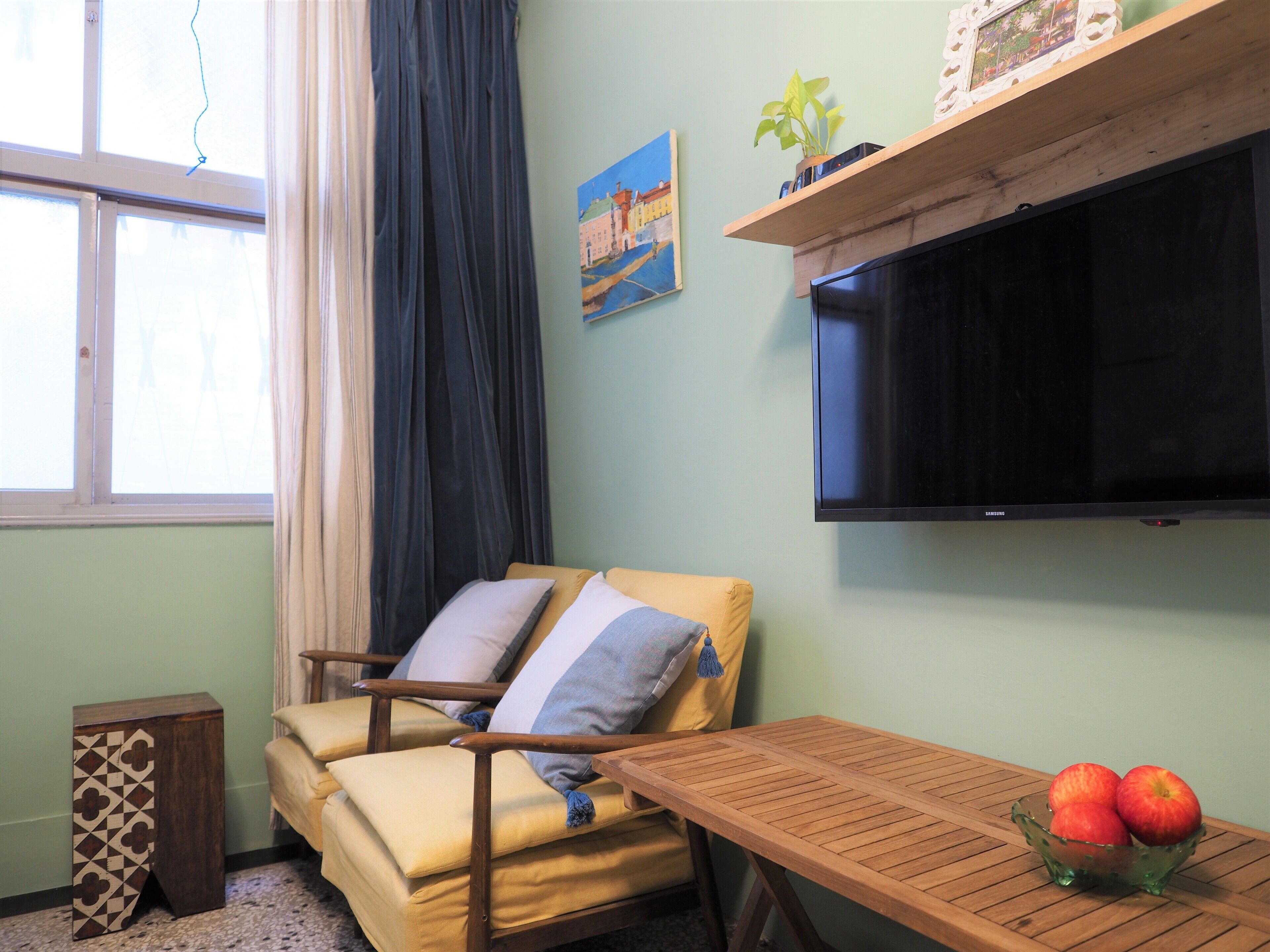 A room with a yellow armchair, a wooden table with a bowl of apples, a flat-screen TV, a painting, and a window with curtains.