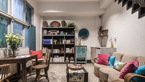 Una sala de estar acogedora con una mesa de madera, sillas de mimbre, un sofá con cojines coloridos, una estantería llena de libros y objetos decorativos, y un ventanal con cortinas.