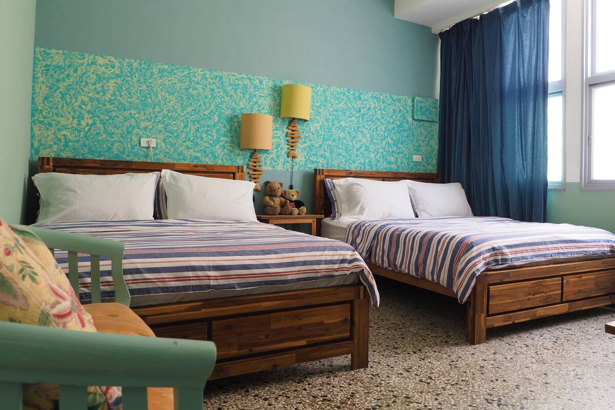 A room with two wooden beds, a nightstand with a lamp, a teddy bear, and a window with blue curtains.