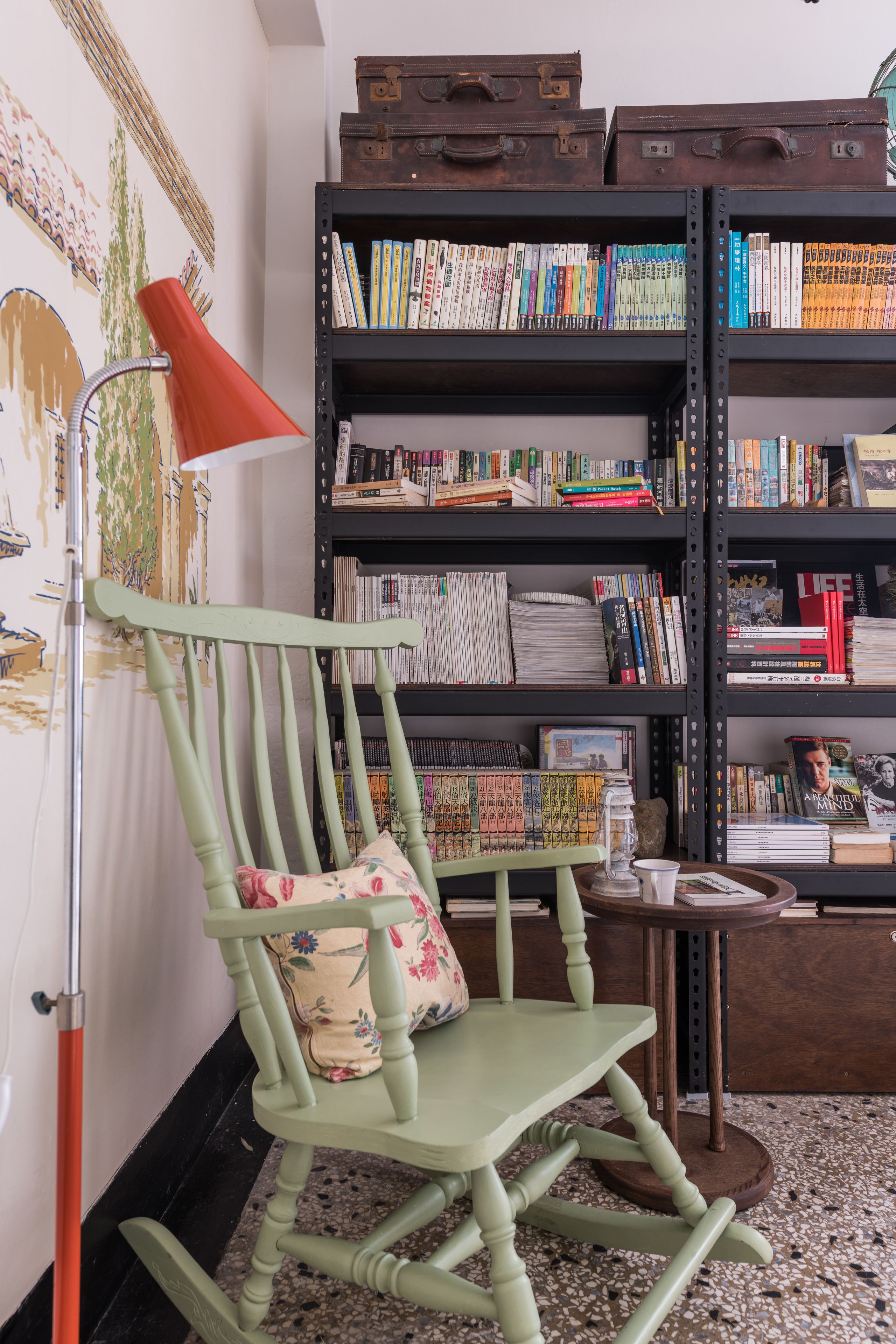 Ein Zimmer mit einem grünen Schaukelstuhl, einem Bücherregal voller Bücher und einem alten Koffer darauf.