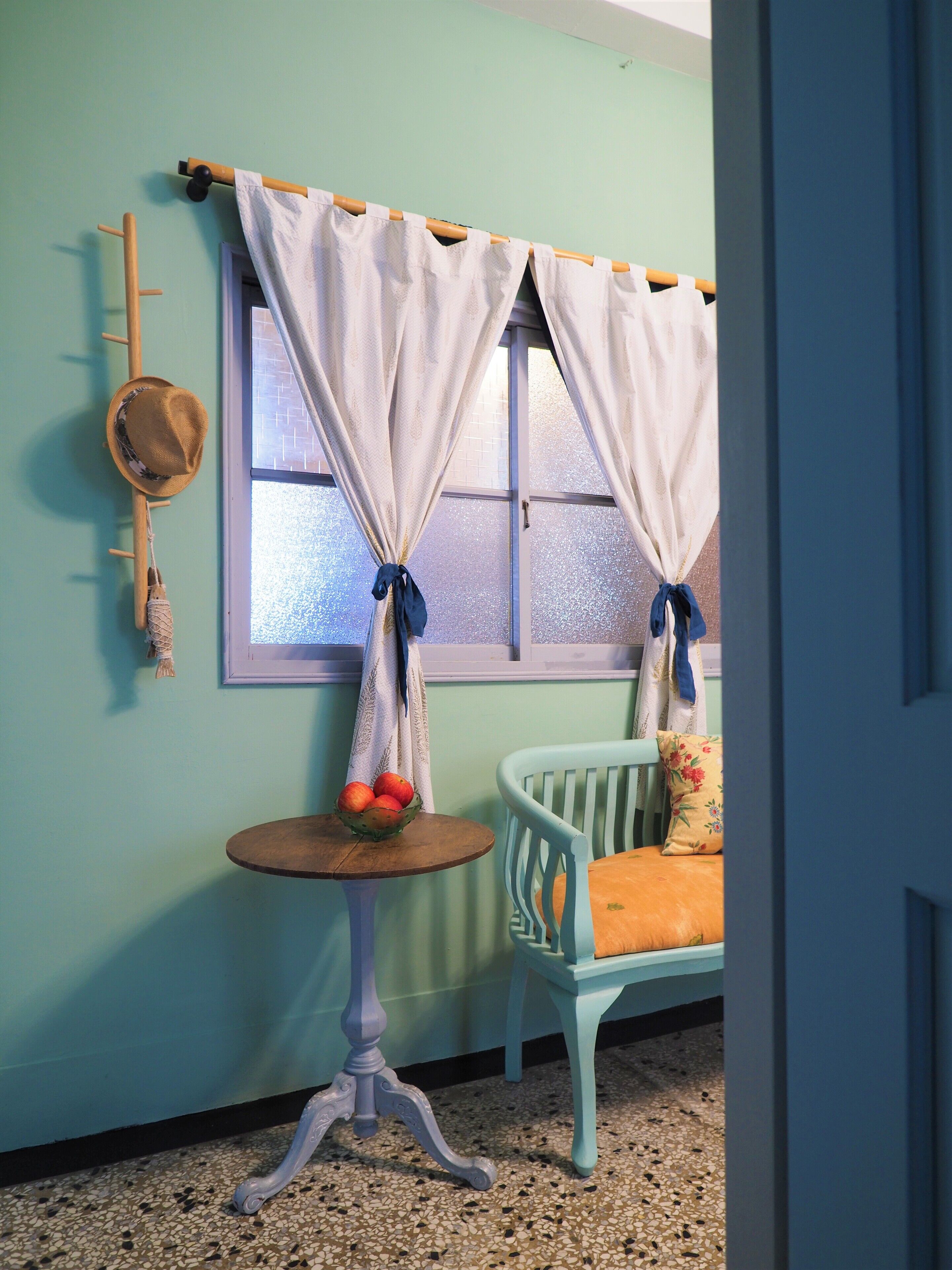 A room with teal walls, a window with curtains, a wooden coat rack, a small round table with a bowl of fruit, and a light blue chair with a cushion.