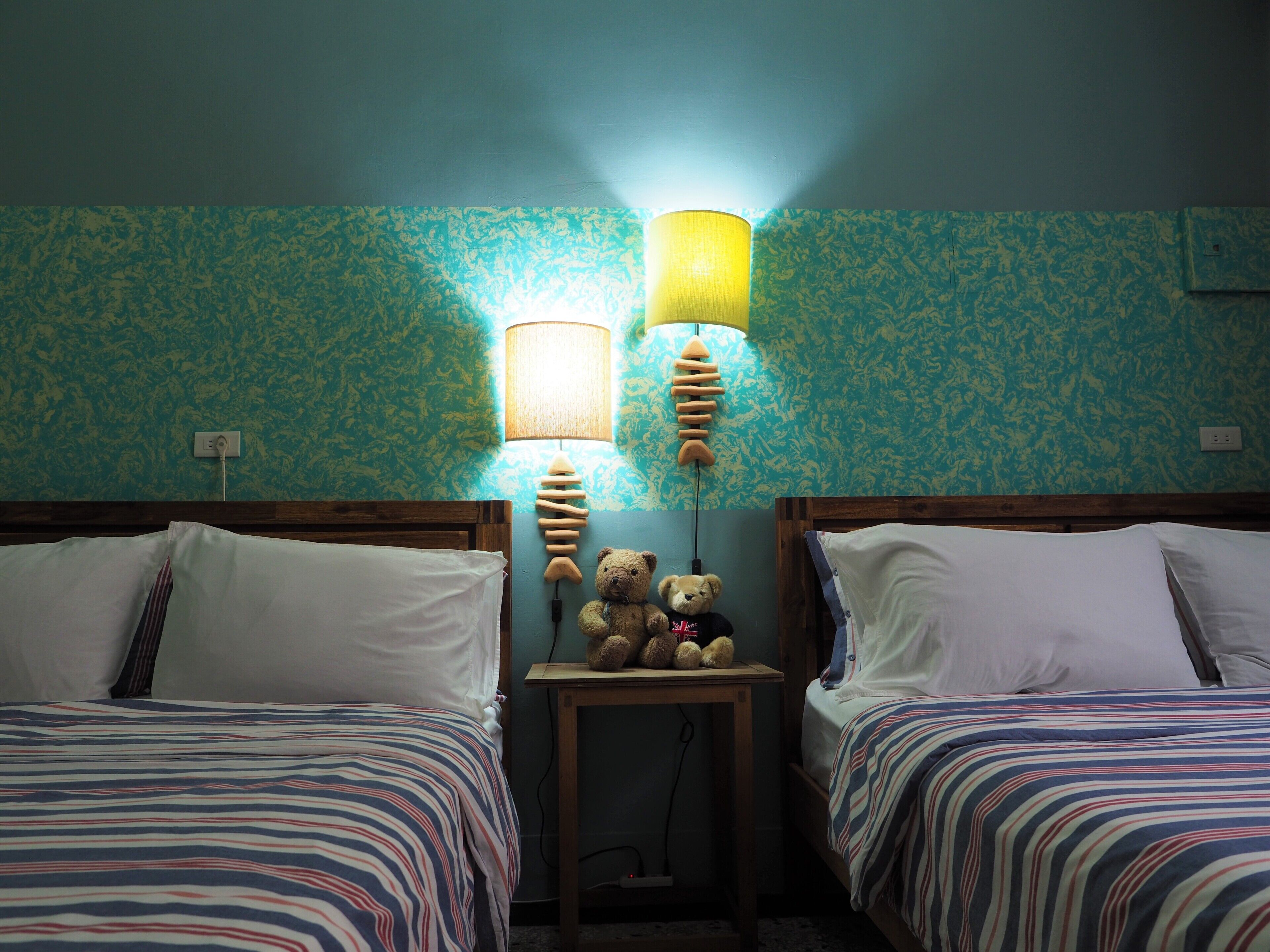 A room with two beds, striped bedding, a small table with a lamp, and a wall with a textured pattern.