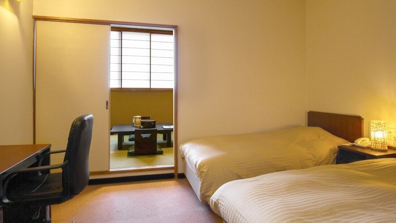 Habitación de hotel con cama, escritorio, silla, mesita con lámpara y puerta corredera que da a un balcón.