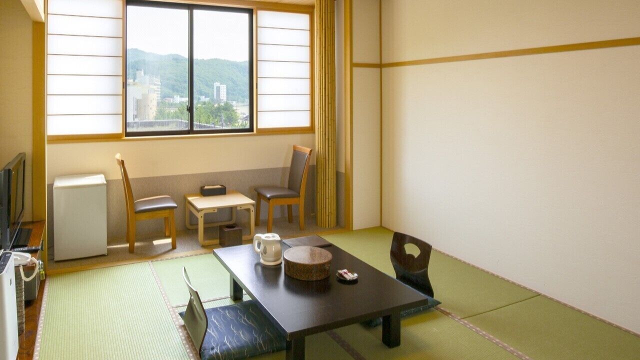 Una habitación japonesa tradicional con suelo de tatami, una mesita baja, sillas, un pequeño calentador y una ventana con vistas a la montaña.