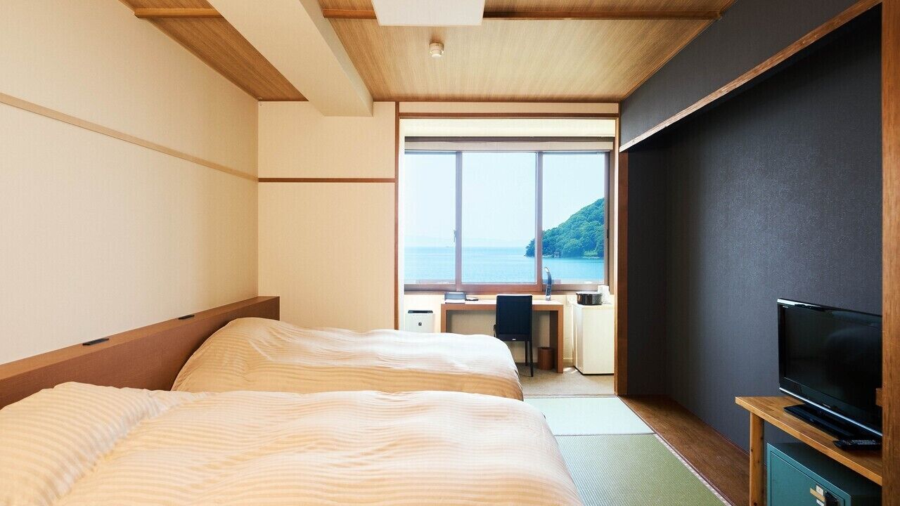 Ein Hotelzimmer mit zwei Betten, einem Schreibtisch, einem Fernseher und einem großen Fenster mit Meerblick.